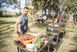 Stono Outdoor Living A man in an apron cooks meat and fresh vegetables on the Tagwood BBQ Height Adjustable Griddle - BBQ54SS with a 304 stainless steel surface, while people enjoy a meal under trees in a sunlit, grassy park.