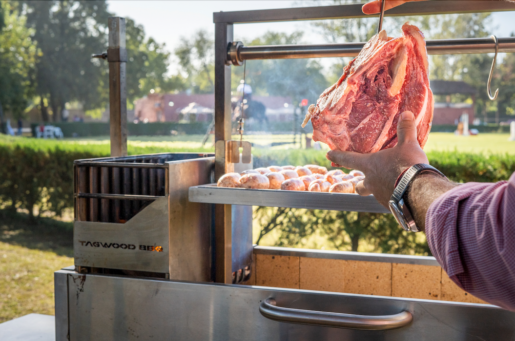Stono Outdoor Living Outdoors in a sunny park, a person uses Tagwood BBQ S Shape Meat Hooks (set of 7) - BBQ56SS to hang meats over a barbecue grill with sausages cooking below.
