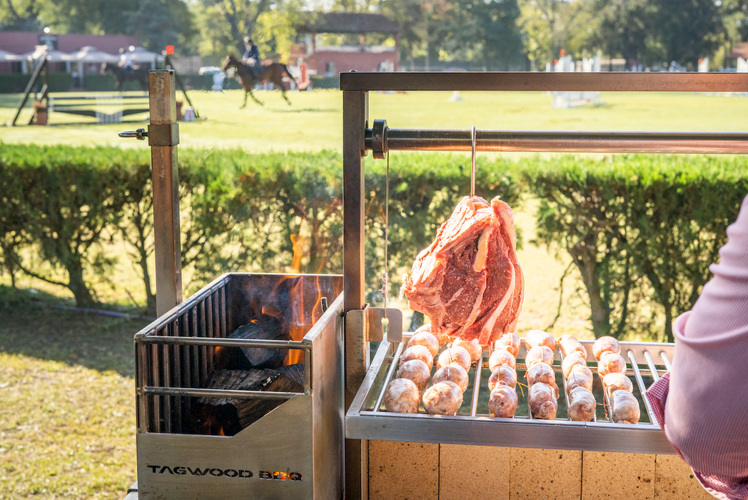 Stono Outdoor Living Outdoors near a grassy field, a Tagwood BBQ S Shape Meat Hooks (set of 7) by Tagwood BBQ holds sausages and a large cut of meat on a 304 stainless steel grill, slow-cooking as someone rides horseback in the background.