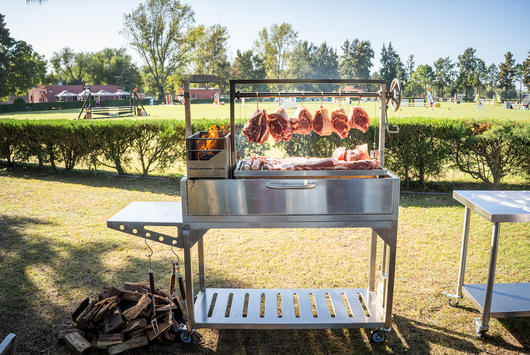 Stono Outdoor Living An outdoor barbecue features meat slabs slow cooking on a 304 stainless steel grill with Tagwood BBQ S Shape Meat Hooks (set of 7, BBQ56SS) by Tagwood BBQ, amid grassy fields, trees, and a white fence. Stacked firewood lies nearby.