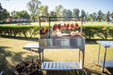 Stono Outdoor Living An outdoor barbecue features meat slabs slow cooking on a 304 stainless steel grill with Tagwood BBQ S Shape Meat Hooks (set of 7, BBQ56SS) by Tagwood BBQ, amid grassy fields, trees, and a white fence. Stacked firewood lies nearby.