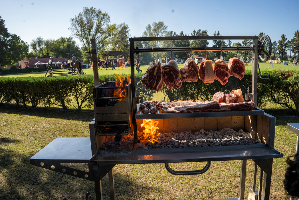 Stono Outdoor Living Tagwood BBQ S Shape Meat Hooks (set of 7) by Tagwood BBQ hold various cuts of meat slow-cooking over an open flame at a grassy park barbecue, with people and trees in the background.