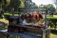 Stono Outdoor Living Several large cuts of meat are slow cooking outdoors on a 304 stainless steel grill, suspended with Tagwood BBQ S Shape Meat Hooks (set of 7) - BBQ56SS, by Tagwood BBQ, with trees, grass, and people visible on a sunny day.