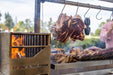 Stono Outdoor Living Large cuts of meat hang above a grill on Tagwood BBQ S Shape Meat Hooks (set of 7, BBQ56SS) by Tagwood BBQ. Outdoors, smoke rises as the fire cooks the meat; a person in a pink shirt stands in the background.