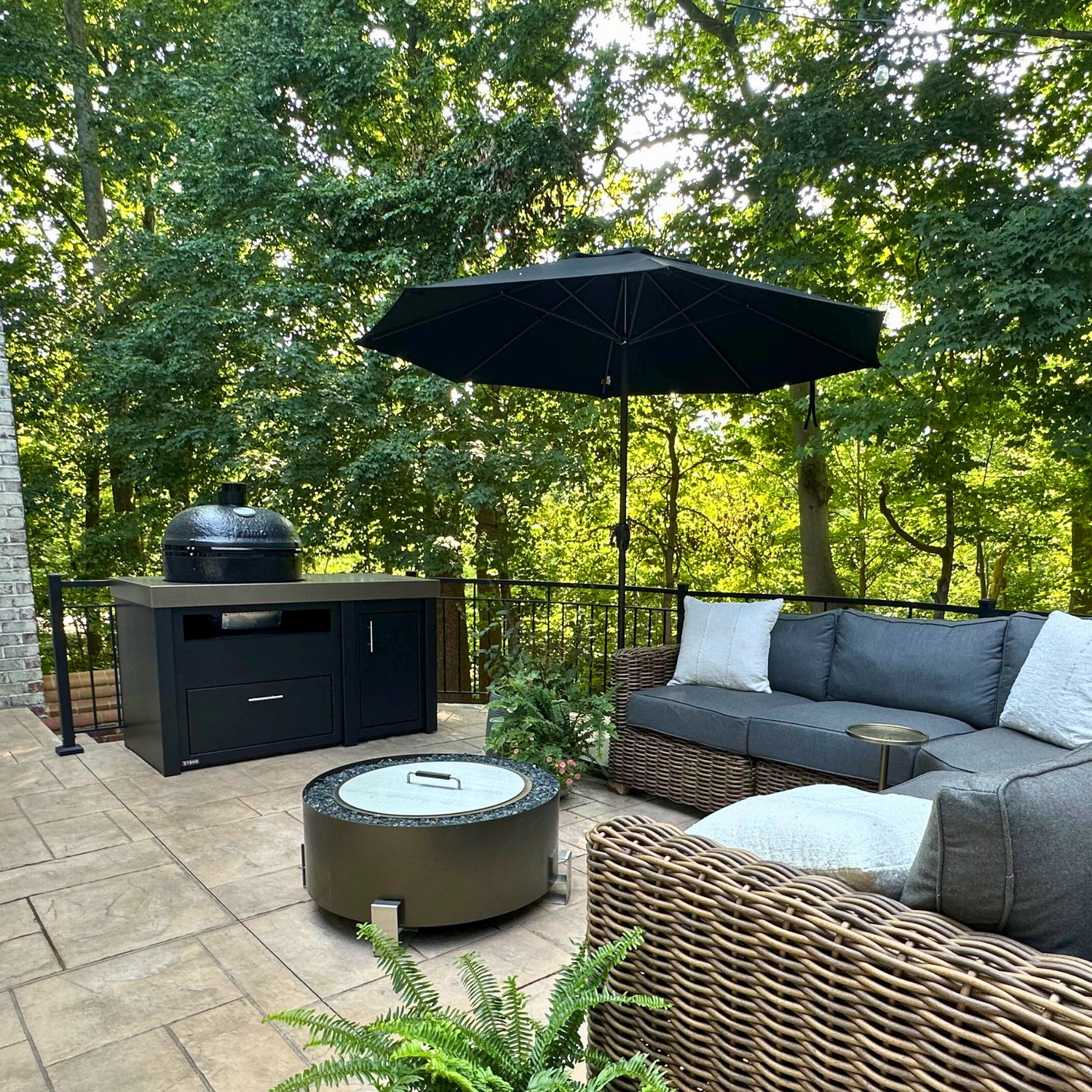 Stono Outdoor Living A cozy outdoor patio with a wicker sectional sofa, a round fire pit, a black grill station, a black umbrella, and surrounding green trees providing shade. The area is paved with stone tiles.