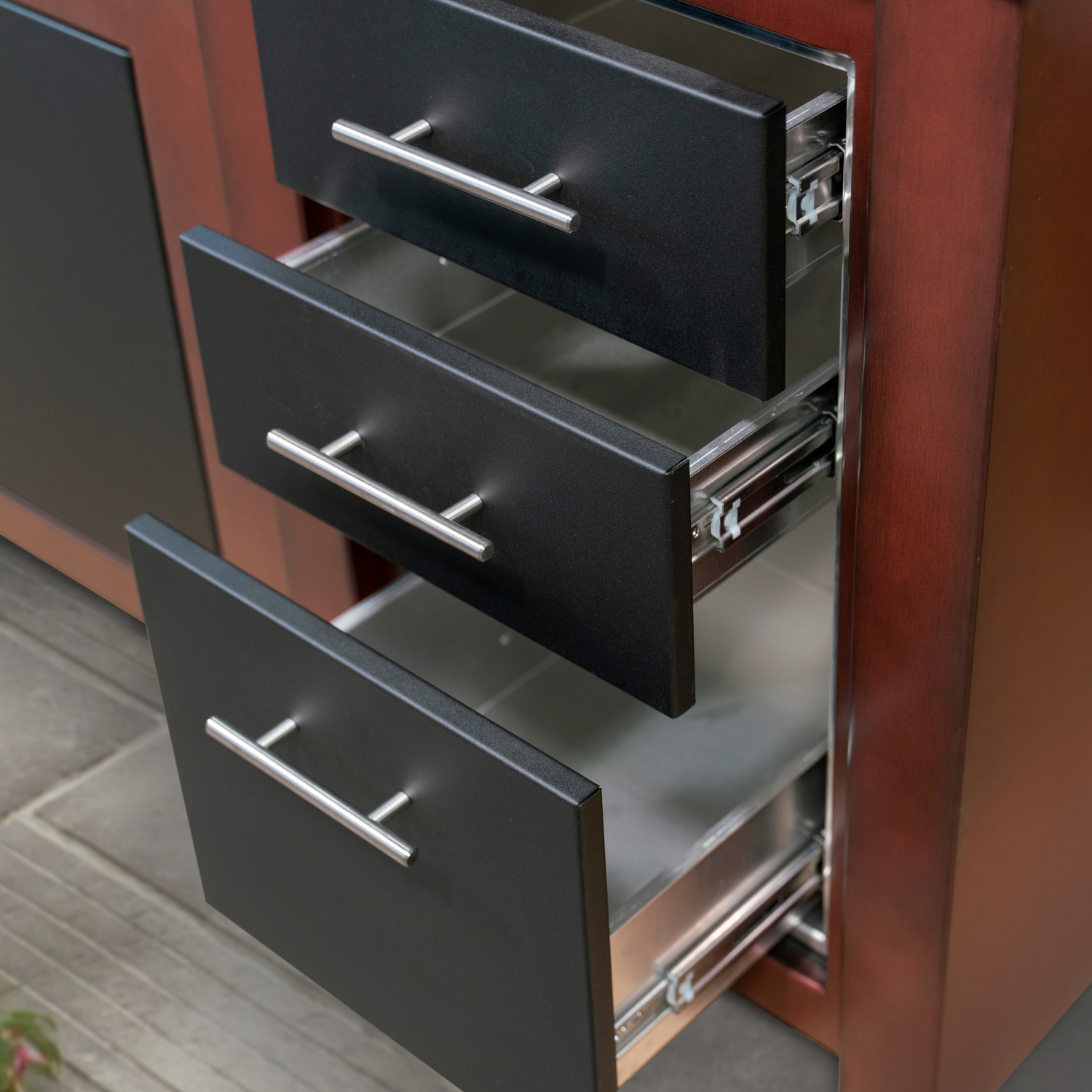 Stono Outdoor Living Three stainless steel drawers with black fronts are open on a brown wooden cabinet, revealing empty interiors. The cabinet is placed on a tiled floor.