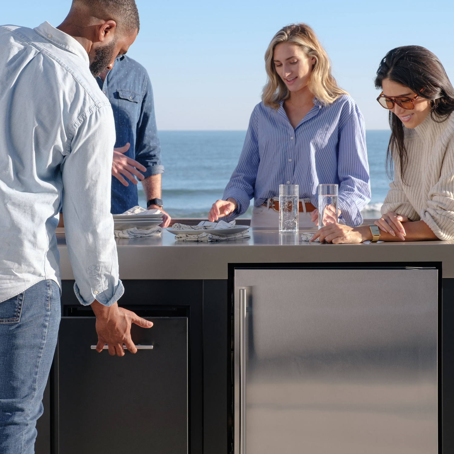 Stono Outdoor Living Four people gather around an outdoor kitchen counter by the ocean, smiling and preparing a meal together under a clear sky. Two glasses of water and plates are on the counter.