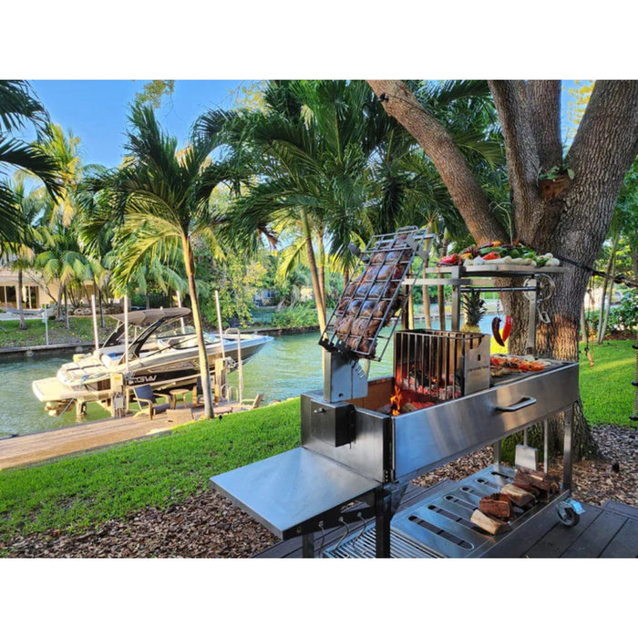 Stono Outdoor Living The Tagwood BBQ Argentine Santa Maria Wood Fire & Charcoal Grill (BBQ06SS) by Tagwood BBQ, filled with skewers and vegetables, stands beside a canal with a docked boat, surrounded by palm trees and greenery on a sunny day.
