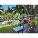 Stono Outdoor Living The Tagwood BBQ Argentine Santa Maria Wood Fire & Charcoal Grill (BBQ06SS) by Tagwood BBQ, filled with skewers and vegetables, stands beside a canal with a docked boat, surrounded by palm trees and greenery on a sunny day.