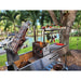 Stono Outdoor Living The Tagwood BBQ Argentine Santa Maria Wood Fire & Charcoal Grill (BBQ06SS) by Tagwood BBQ, made of 304 stainless steel, is set up beside a canal with boats and palm trees, wood-fired skewers grilling meats and vegetables to perfection.