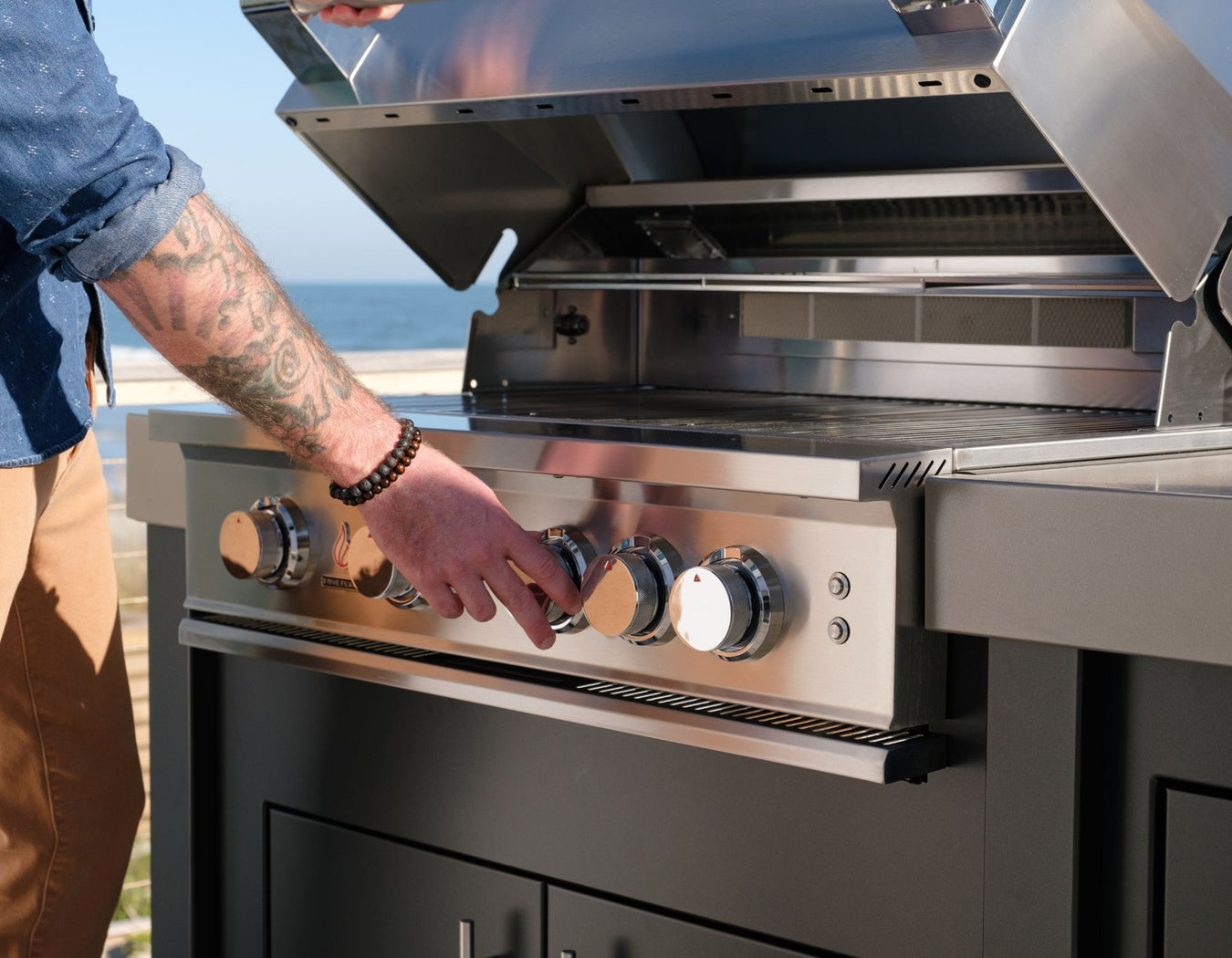 Stono Outdoor Living A person with tattooed arms and a beaded bracelet adjusts the knobs on a stainless steel outdoor grill with the lid open, near a beach or coastal area.