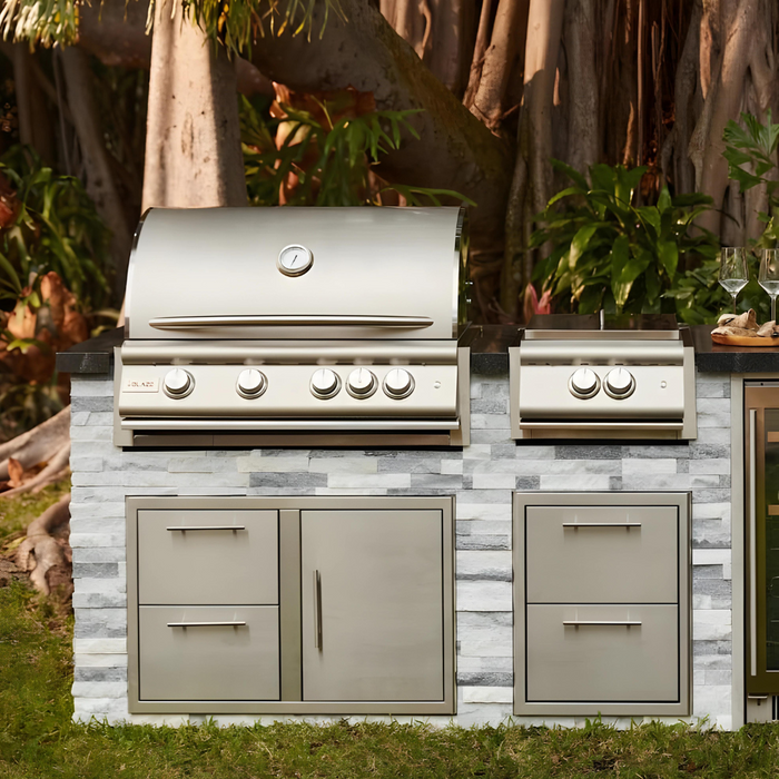 Stono Outdoor Living The Blaze Premium LTE+ 32" 4-Burner Propane Grill with rear infrared burner and grill lights by Blaze is built into a stone counter with storage drawers, set among green plants and trees in a tranquil garden.