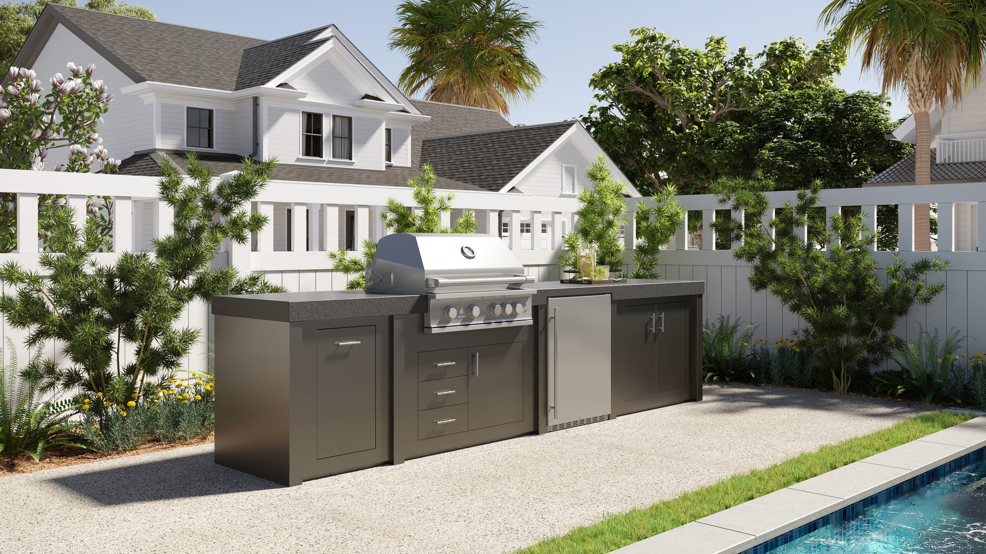 Stono Outdoor Living Outdoor kitchen with a built-in stainless steel grill, cabinets, and countertop, set on a patio beside a swimming pool, with white fencing, green plants, and houses in the background on a sunny day.