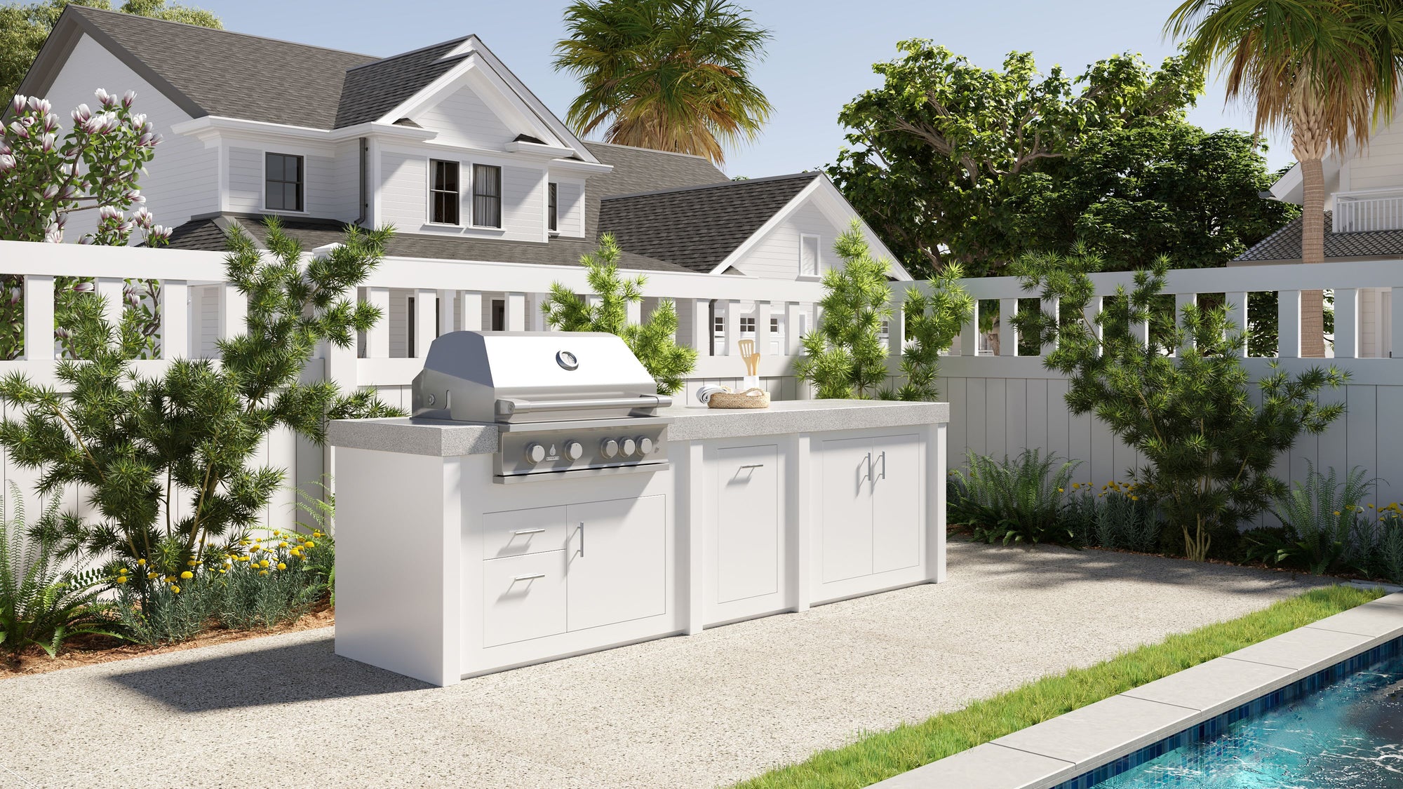 Stono Outdoor Living A modern outdoor kitchen with a stainless steel grill and white cabinets sits on a patio near a swimming pool, surrounded by greenery and a white fence, with houses and trees in the background.