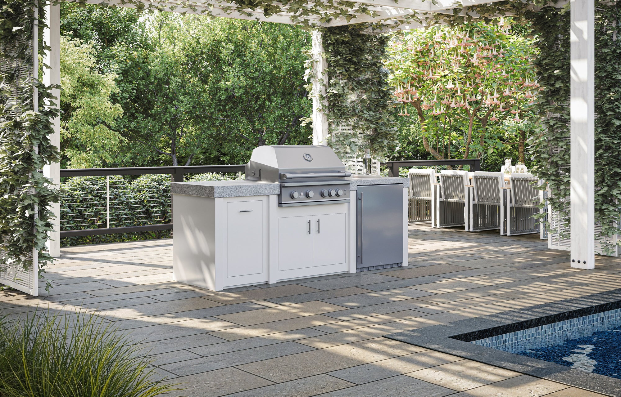 Stono Outdoor Living Outdoor kitchen with a modern grill, cabinets, and mini fridge under a pergola covered in greenery. A dining table with chairs sits nearby, next to a pool and surrounded by lush trees and plants.