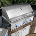 Stono Outdoor Living A Delta Heat 32-inch 3-burner built-in propane gas grill (DHBQ32G-DL) features three blue-lit knobs and a closed lid, set in a stone and granite outdoor kitchen surrounded by greenery.