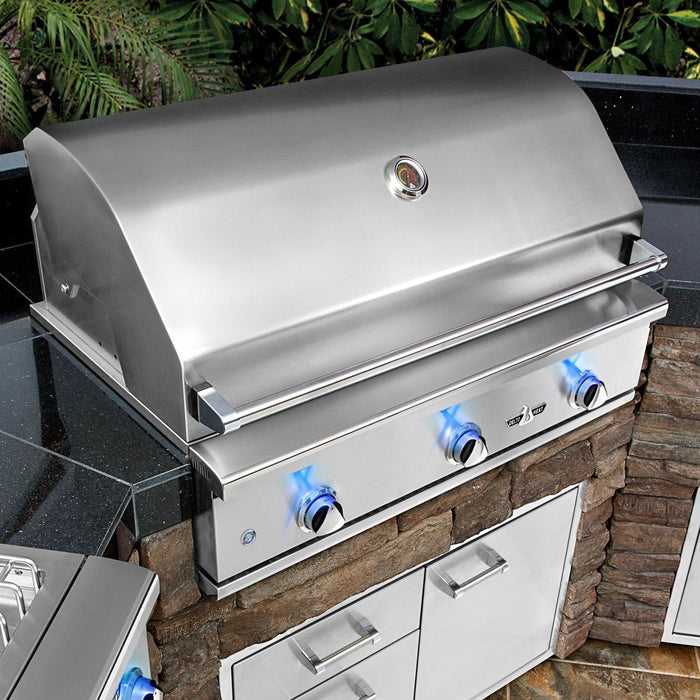 Stono Outdoor Living The Delta Heat 38-Inch 3-Burner Built-In Propane Gas Grill (DHBQ38G-DL) features three blue-lit knobs on a stone counter with built-in cabinets, forming a sleek outdoor kitchen framed by lush greenery.