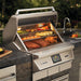 Stono Outdoor Living A Twin Eagles Wi-Fi Controlled 36-Inch Built-In Stainless Steel Pellet Grill and Smoker (TEPG36R) with rotisserie is built into a stone countertop with a digital control panel, surrounded by greenery and a nearby sink.