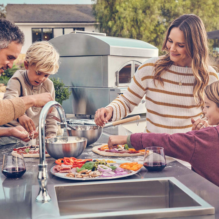 Stono Outdoor Living A family enjoys making artisan pizza together on the Delta Heat 30-Inch Built-In Pizza Oven (DHPO30BI-N). Fresh ingredients and red wine complete the warm, cheerful atmosphere around the high-quality outdoor kitchen centerpiece.