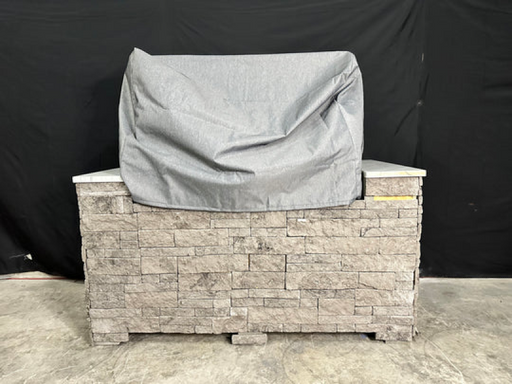 Stono Outdoor Living A stone outdoor kitchen island with a marble countertop is protected by the Tagwood BBQ BBQ05SS Cover - COVE05, a gray, water-resistant BBQ cover from Tagwood BBQ, set against a black backdrop on a concrete floor.