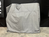 Stono Outdoor Living A large object covered with a gray, UV-resistant Tagwood BBQ BBQ23SS Cover (COVE23) stands on a concrete floor before a black backdrop. A small Tagwood BBQ label appears on the bottom right corner of the water-resistant cover.