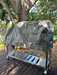 Stono Outdoor Living A Tagwood BBQ outdoors metal grill, protected by the Tagwood BBQ BBQ08SS Cover - COVE08, stands on a wooden deck beneath a large tree amid greenery and sunlight.