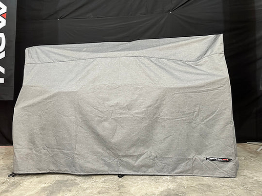 Stono Outdoor Living A large rectangular object is covered with a gray, water-resistant Tagwood BBQ BBQ24SS Cover - COVE24, placed indoors on a concrete floor. A small Tagwood BBQ branded label appears on the lower right corner of the cover.