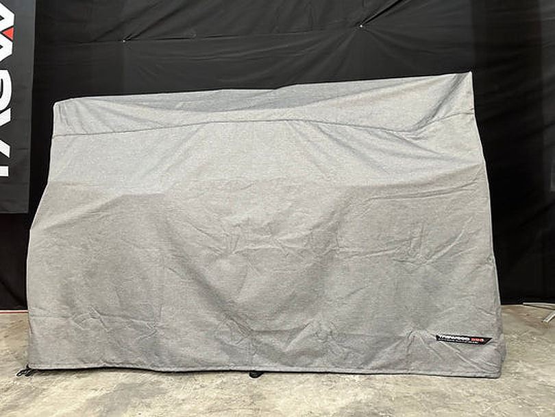 Stono Outdoor Living A large rectangular object is covered with a gray, water-resistant Tagwood BBQ BBQ24SS Cover - COVE24, placed indoors on a concrete floor. A small Tagwood BBQ branded label appears on the lower right corner of the cover.