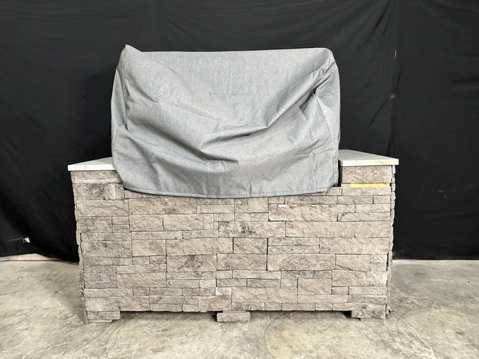 Stono Outdoor Living A stone outdoor kitchen island with a covered grill sits on concrete, protected by the Tagwood BBQ BBQ25SS Cover - COVE25 from Tagwood BBQ. A black fabric backdrop completes the scene.
