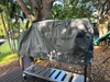 Stono Outdoor Living The Tagwood BBQ BBQ28SS Cover - COVE28 protects a grill on a wooden deck, surrounded by greenery. Sunlight filters through the trees, casting dappled shadows on this UV-resistant cover from Tagwood BBQ.