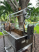 Stono Outdoor Living The Tagwood BBQ Stainless Steel Rotisserie Kit - BBQ50SS PRO by Tagwood BBQ is set up on a deck near trees and a canal, with a wheel, electric motor, lush greenery in the background, and a dog visible.