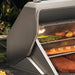 Stono Outdoor Living A close-up of the Twin Eagles Wi-Fi Controlled 36-Inch Built-In Stainless Steel Pellet Grill and Smoker (TEPG36G), featuring shelves with vegetables, fish, and meat amid lush garden greenery.