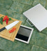 Stono Outdoor Living A Twin Eagles Trash Chute with Cutting Board (TETC) sits on green tiled flooring, with sliced tomatoes, chili peppers, and a knife on the board above a floor compartment with a mirrored interior and a removed cover.